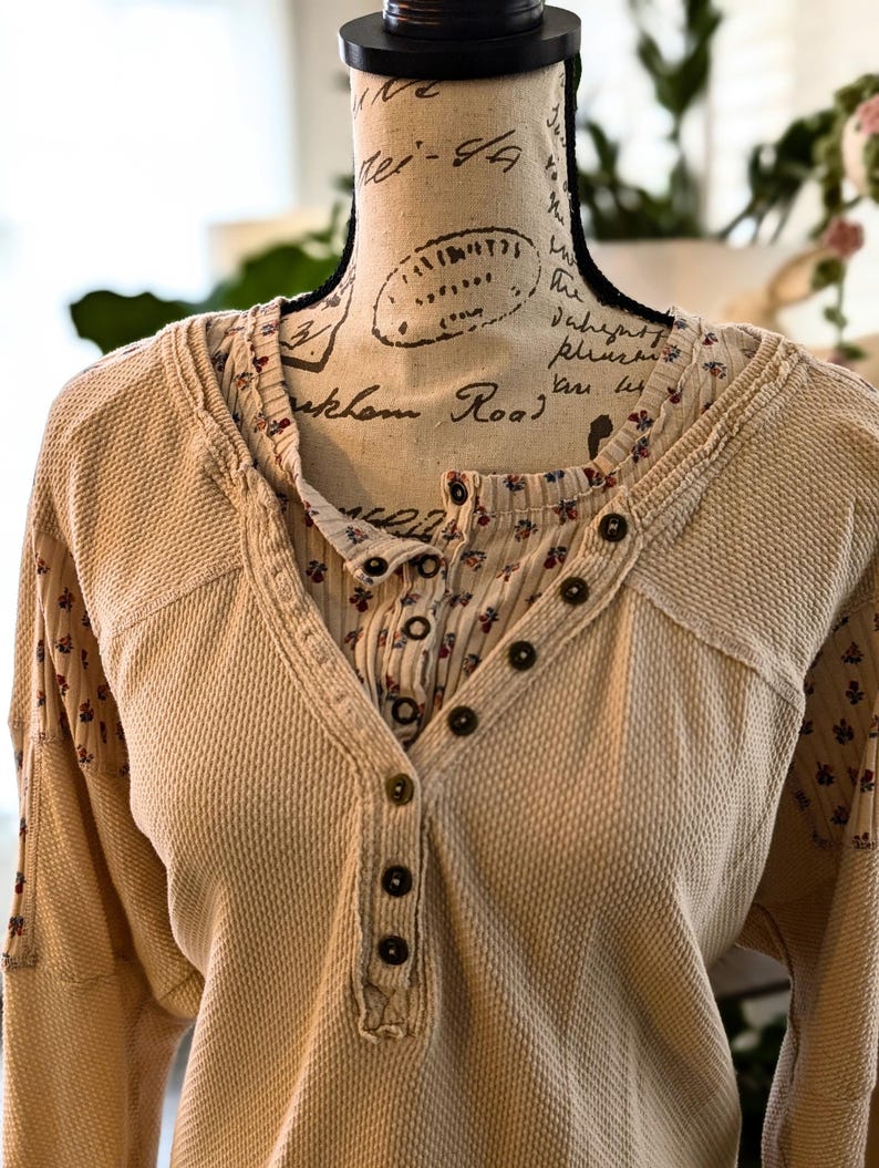 Puede incluir: Una camisa henley beige de punto de gofre con una capa interior y mangas con estampado floral. La camisa tiene una parte delantera abotonada y mangas largas. La cabeza del maniqu&iacute; es visible en el fondo.