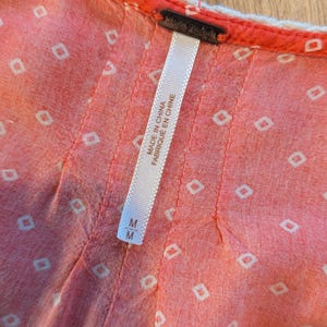 May include: Close-up of a coral-colored garment with a white geometric pattern. The garment has a white trim and a tag that reads "Free People" and "Made in China Fabriqu&eacute; en Chine". The size is M.