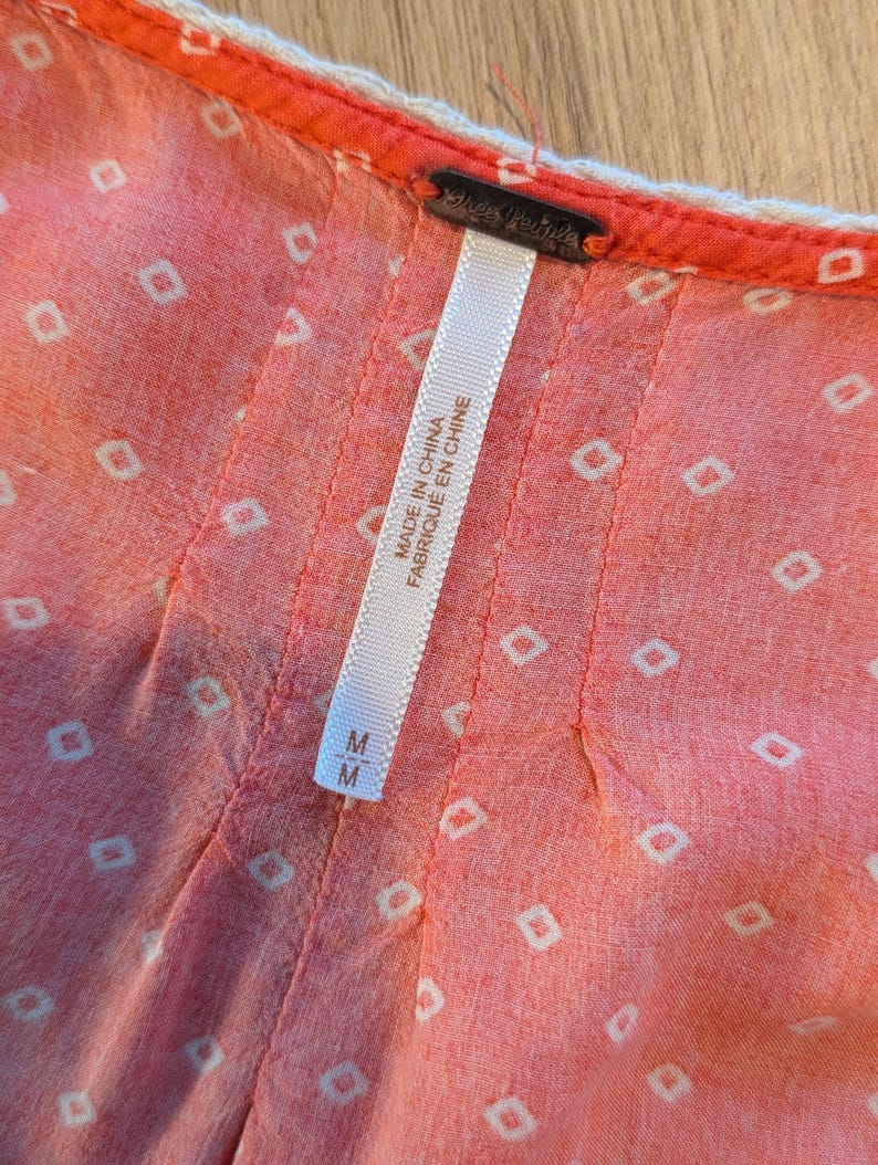 May include: Close-up of a coral-colored garment with a white geometric pattern. The garment has a white trim and a tag that reads "Free People" and "Made in China Fabriqu&eacute; en Chine". The size is M.