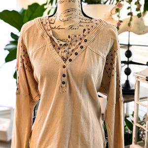 Puede incluir: Camisa beige de manga larga de punto de gofre con cuello en V y cierre de botones. La camisa presenta tela estampada floral en el cuello interior y los pu&ntilde;os. La camisa se exhibe en un maniqu&iacute;.