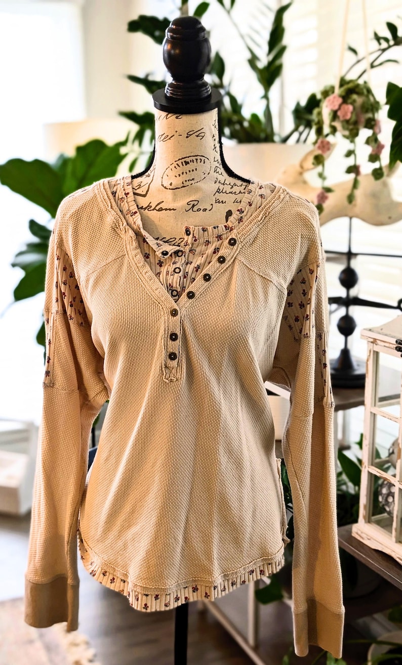 Puede incluir: Camisa beige de manga larga de punto de gofre con cuello en V y cierre de botones. La camisa presenta tela estampada floral en el cuello interior y los pu&ntilde;os. La camisa se exhibe en un maniqu&iacute;.