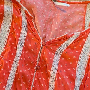 May include: Close-up of a coral-colored top with a V-neck and button-down front. The fabric features a white dotted pattern and is trimmed with white lace. The top is a casual, feminine garment.