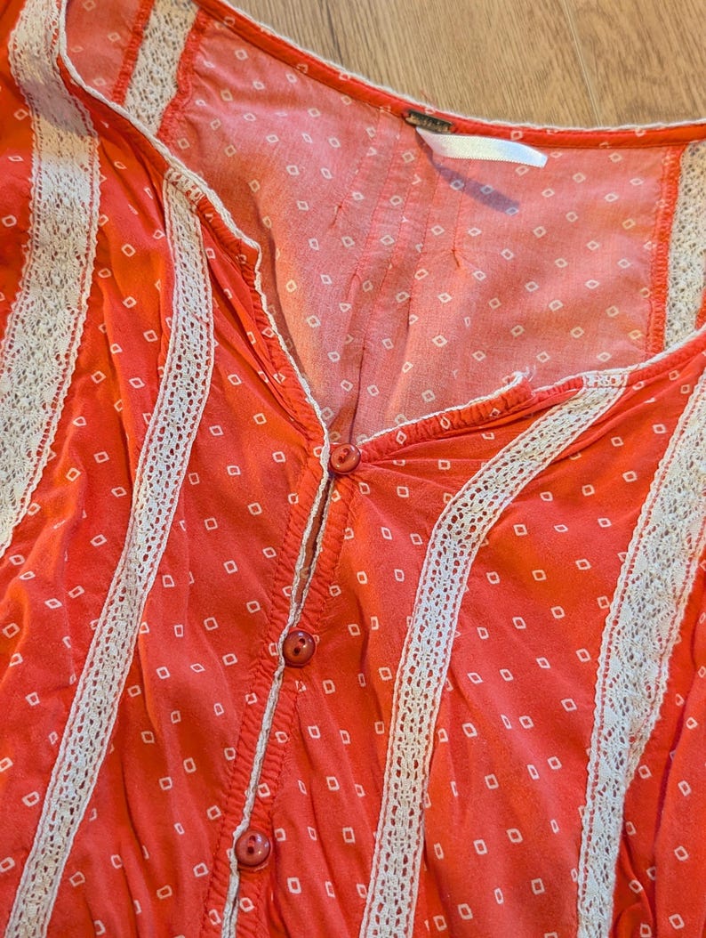 May include: Close-up of a coral-colored top with a V-neck and button-down front. The fabric features a white dotted pattern and is trimmed with white lace. The top is a casual, feminine garment.
