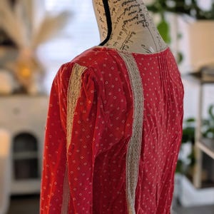 May include: A red long-sleeved blouse with a small white diamond pattern. The blouse features beige lace trim along the sleeves and front. The garment is displayed on a mannequin.