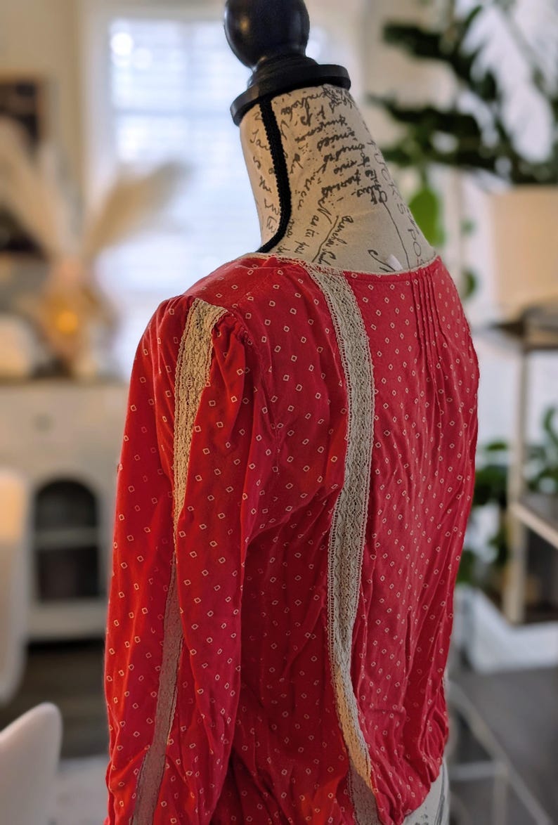 May include: A red long-sleeved blouse with a small white diamond pattern. The blouse features beige lace trim along the sleeves and front. The garment is displayed on a mannequin.