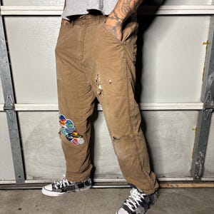 K&ouml;nnte beinhalten: Braune Arbeitshose mit Used-Details und einem bunten Aufn&auml;her am linken Knie. Die Hose wird mit schwarz-wei&szlig; gemusterten Turnschuhen kombiniert. Die Person tr&auml;gt ein graues T-Shirt.