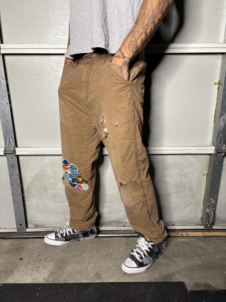 K&ouml;nnte beinhalten: Braune Arbeitshose mit Used-Details und einem bunten Aufn&auml;her am linken Knie. Die Hose wird mit schwarz-wei&szlig; gemusterten Turnschuhen kombiniert. Die Person tr&auml;gt ein graues T-Shirt.