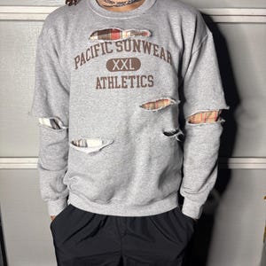 May include: Gray sweatshirt with distressed details and plaid fabric peeking through the rips. The front features the text "PACIFIC SUNWEAR XXL ATHLETICS" in brown lettering. The sweatshirt is paired with black pants.