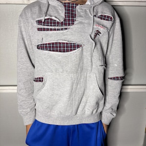 May include: A gray hoodie with distressed details, featuring red and black plaid fabric peeking through the cutouts. The hoodie has a front pocket and a hood. The Atlanta Falcons logo is visible on the right side.