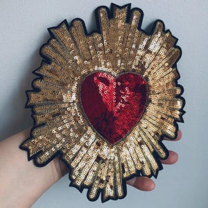 Peut inclure: Un écusson en forme de cœur avec un centre rouge à paillettes, entouré de paillettes dorées et d'une bordure noire. Le patch présente un motif de rayons de soleil, créant un effet radiant. Ce patch décoratif peut être utilisé pour les vêtements ou les accessoires.