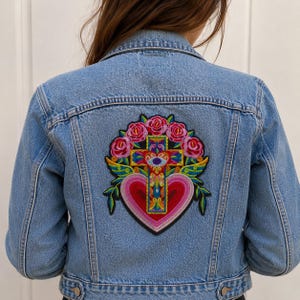 Peut inclure: Une veste en jean bleu clair avec un grand écusson brodé coloré au dos. L'écusson représente une croix centrale avec un motif d'œil, entourée de roses roses, de feuilles vertes et d'un grand cœur rouge à la base avec des détails complexes.