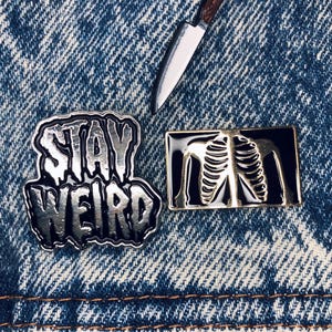 Peut inclure: Épingles en émail et un petit couteau sur fond denim. Les épingles comprennent une épingle argentée avec l'inscription « STAY WEIRD », une épingle squelette noire et dorée et un petit couteau avec un manche marron.