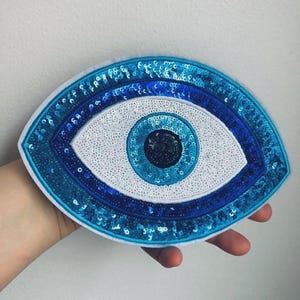 Peut inclure: Un écusson décoratif en forme d'œil, avec un centre blanc et des cercles concentriques de paillettes bleues. Le bord extérieur est bleu foncé, créant un contraste saisissant. Conçu pour la décoration, il mesure environ 10 cm de long.