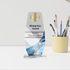 May include: A clear acrylic Rising Star Award with a gold emblem and the text "Rising Star Award" and "Awarded to Emily Chen." The award features a blue and white abstract design and sits on a clear base. Pens and pencils are in a yellow cup in the background.