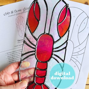 Op de afbeelding: Een gebrandschilderde glazen zonnevanger in de vorm van een kreeft met rode, roze en witte segmenten, omlijnd in zwart. Het ontwerp wordt door een hand vastgehouden, met een patroon en de woorden "digitale download" op de achtergrond.