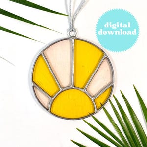 May include: A stained glass sun catcher with a yellow and white design. The sun catcher is round and has a silver frame. It is hanging from a silver cord.