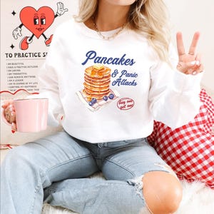 May include: White sweatshirt with a graphic of a stack of pancakes and blueberries with the text "Pancakes & Panic Attacks" and "buy one get one" in red.