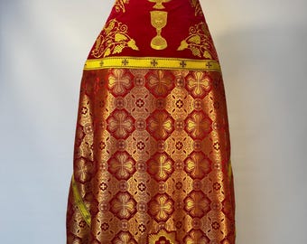 May include: A red and gold liturgical vestment featuring a repeating cross pattern in metallic gold thread. The upper section displays an embroidered gold chalice and a dove symbol, bordered by yellow bands with small cross motifs throughout.