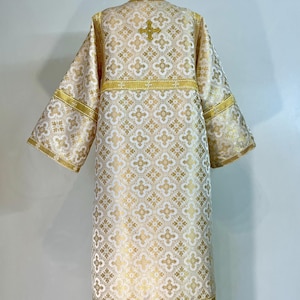 May include: White liturgical vestment with gold embroidery of a cross and geometric patterns. The vestment has long sleeves and a wide, flowing skirt.