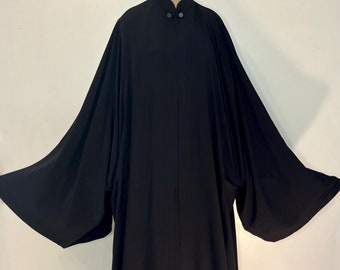 Puede incluir: Una larga túnica negra con cuello mandarín y dos botones. La túnica tiene mangas anchas y una falda larga y fluida.