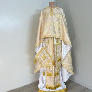 May include: A white and gold liturgical vestment with intricate embroidery and a matching stole. The vestment is worn by a priest during religious ceremonies.