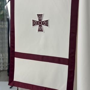 May include: A white liturgical banner with a burgundy border and accents. The banner features an embroidered cross in the center and a horizontal burgundy stripe. The design is simple and elegant, suitable for religious ceremonies.