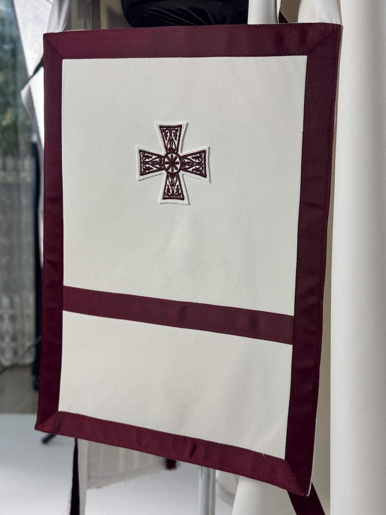 May include: A white liturgical banner with a burgundy border and accents. The banner features an embroidered cross in the center and a horizontal burgundy stripe. The design is simple and elegant, suitable for religious ceremonies.