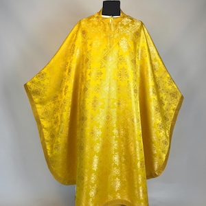 Pode incluir: Uma casula litúrgica amarelo-brilhante com um padrão de damasco repetitivo num tom dourado mais claro. A peça tem uma gola alta, uma silhueta larga e fluida, e é rematada com um debrum dourado a condizer ao longo da bainha curva.
