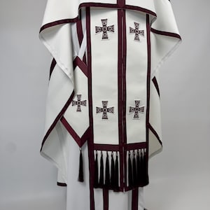 May include: White liturgical vestment with burgundy trim and cross details. The vestment features a cape-like top and a long, flowing design. The front panel has multiple embroidered crosses and a row of black tassels.