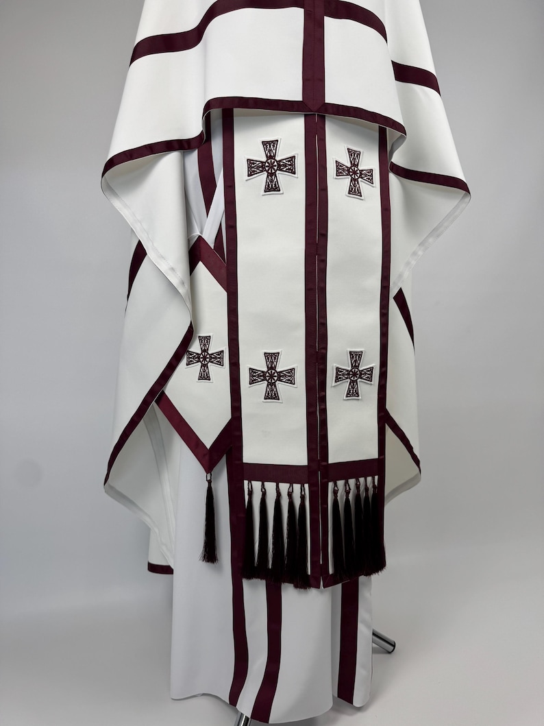 May include: White liturgical vestment with burgundy trim and cross details. The vestment features a cape-like top and a long, flowing design. The front panel has multiple embroidered crosses and a row of black tassels.