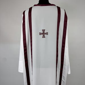 May include: White liturgical vestment with burgundy accents. The back features a burgundy cross design. The vestment has long sleeves and is displayed on a black mannequin head. The fabric appears to be smooth and opaque.