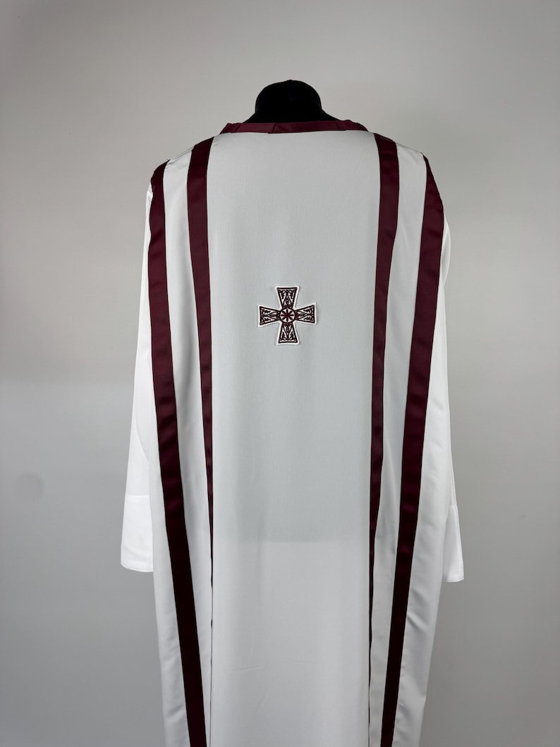 May include: White liturgical vestment with burgundy accents. The back features a burgundy cross design. The vestment has long sleeves and is displayed on a black mannequin head. The fabric appears to be smooth and opaque.