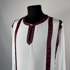 May include: White tunic with long sleeves and vertical burgundy stripes. The tunic has a mandarin collar and a small opening at the neckline, fastened with a single button. The garment is displayed on a black mannequin.
