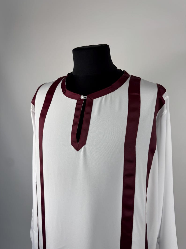 May include: White tunic with long sleeves and vertical burgundy stripes. The tunic has a mandarin collar and a small opening at the neckline, fastened with a single button. The garment is displayed on a black mannequin.