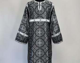 Könnte beinhalten: Ein schwarzes und silbernes liturgisches Gewand mit einem sich wiederholenden kreisförmigen Muster und komplexen Kreuzmotiven. Das Kleidungsstück hat lange, weite Ärmel, einen Rundhalsausschnitt mit vertikalem Schlitz und weiße Zierbänder.