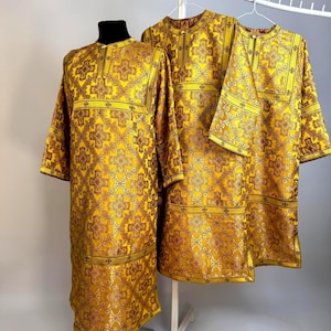 May include: Three long-sleeved tunics in a vibrant gold fabric featuring a repeating dark red and gold cross pattern. Each garment has a high, round neckline and a straight-cut silhouette, with one tunic displayed on a mannequin and two on hangers.