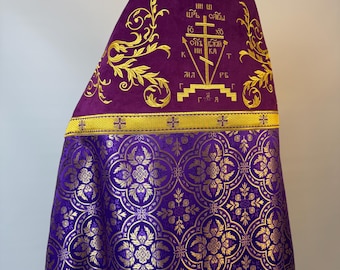 May include: A purple liturgical vestment featuring gold embroidery. The upper section displays a cross with Cyrillic text, while the lower section has a repeating gold floral pattern. A horizontal gold band separates the two distinct fabric sections.