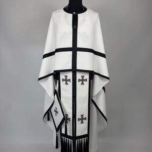 May include: A white liturgical vestment with black trim and crosses. The garment features a high collar, a cape-like design, and a central panel adorned with embroidered crosses. Black tassels hang from the bottom of the panel.