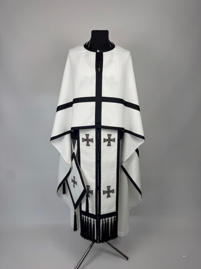 May include: A white liturgical vestment with black trim and crosses. The garment features a high collar, a cape-like design, and a central panel adorned with embroidered crosses. Black tassels hang from the bottom of the panel.