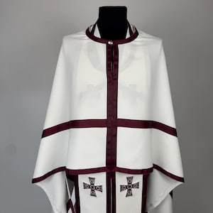 May include: White religious vestment with burgundy trim and cross details. The vestment features a cape-like design with a high collar and a central burgundy stripe. Two embroidered crosses are visible on the lower portion of the garment.