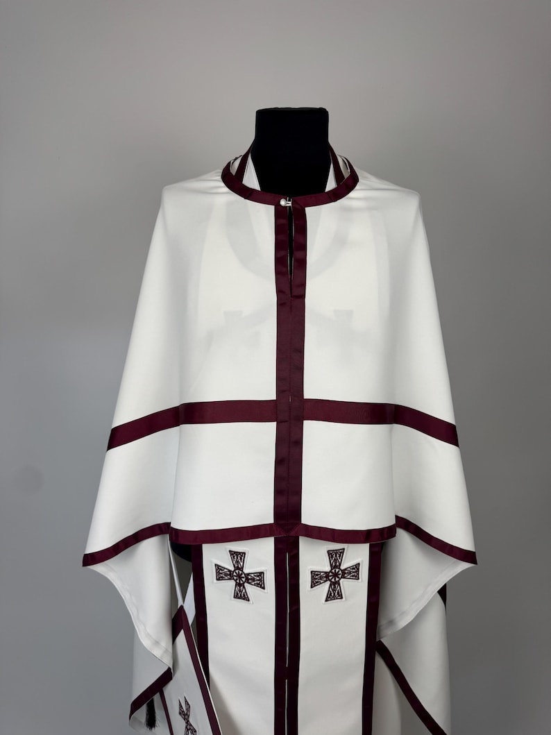 May include: White religious vestment with burgundy trim and cross details. The vestment features a cape-like design with a high collar and a central burgundy stripe. Two embroidered crosses are visible on the lower portion of the garment.