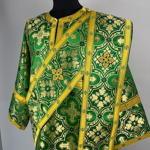 May include: A green and gold liturgical vestment featuring an intricate brocade pattern of floral motifs and crosses. The garment includes a diagonal stole with matching gold trim, a high collar, and wide sleeves, displayed on a black mannequin.