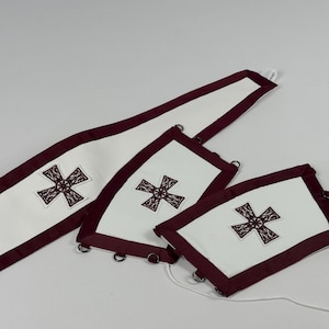 May include: White fabric pieces with a burgundy border and embroidered cross design. The set includes a long, rectangular piece and two smaller, square-shaped pieces. Metal rings are attached to the edges, and a white cord is visible.