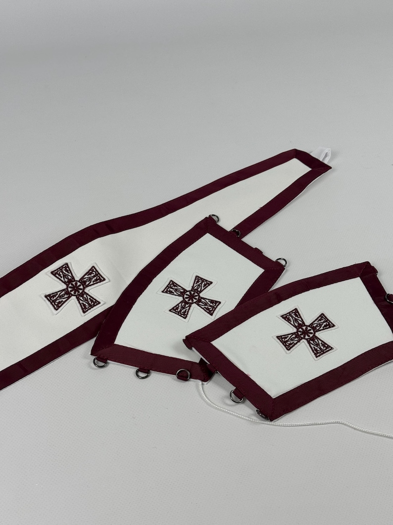 May include: White fabric pieces with a burgundy border and embroidered cross design. The set includes a long, rectangular piece and two smaller, square-shaped pieces. Metal rings are attached to the edges, and a white cord is visible.