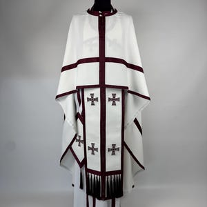 May include: White religious vestment with burgundy trim and cross details. The garment features a high collar, a cape-like top, and a long, flowing skirt. The front panel has embroidered crosses and decorative tassels.