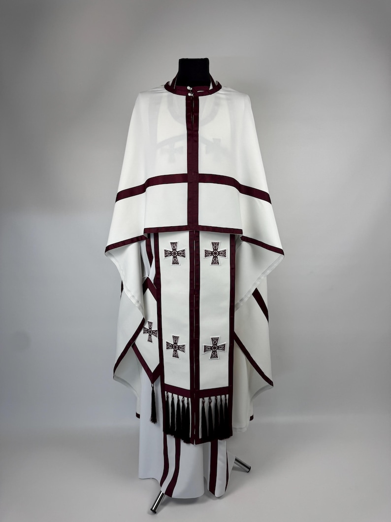 May include: White religious vestment with burgundy trim and cross details. The garment features a high collar, a cape-like top, and a long, flowing skirt. The front panel has embroidered crosses and decorative tassels.