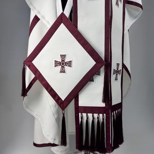 May include: White religious vestment with burgundy trim and embroidered crosses. The set includes a robe, stole, and a square element with tassels. The design features a cross motif and is displayed on a silver stand.