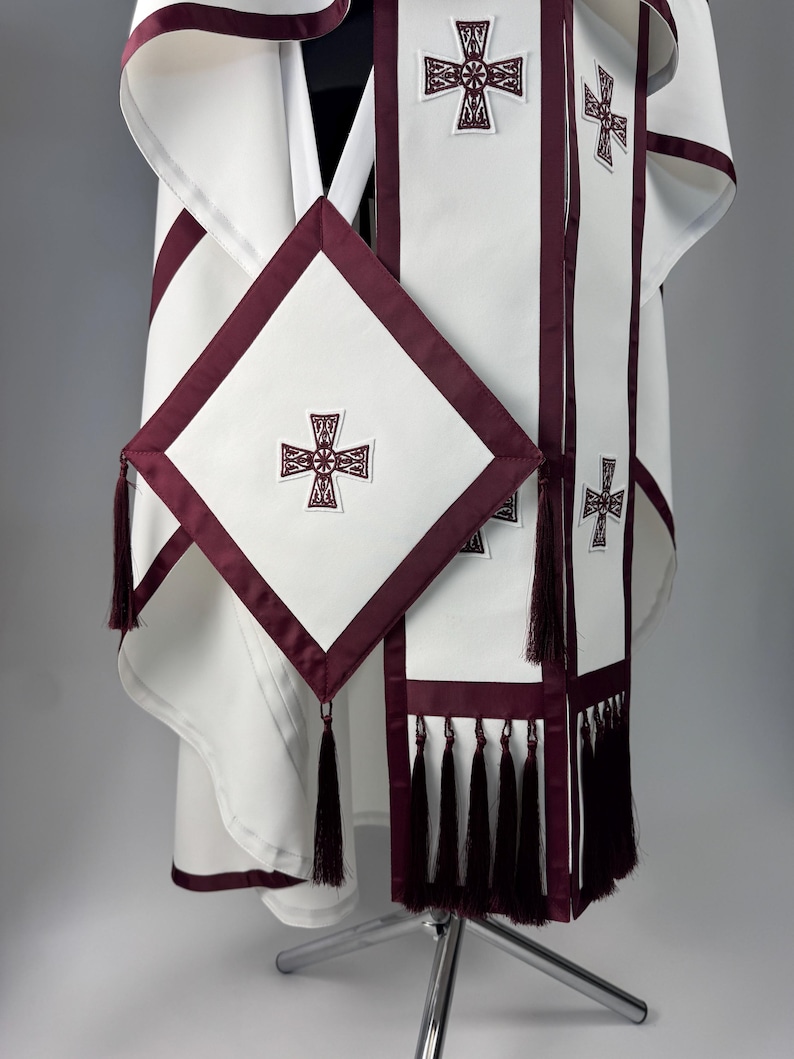 May include: White religious vestment with burgundy trim and embroidered crosses. The set includes a robe, stole, and a square element with tassels. The design features a cross motif and is displayed on a silver stand.