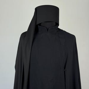 Può includere: Un indumento liturgico nero a tinta unita composto da una tunica a collo alto con chiusura a bottone laterale e un copricapo cilindrico alto abbinato, con un lungo pannello di tessuto drappeggiato che scende lateralmente, creando un aspetto uniforme.