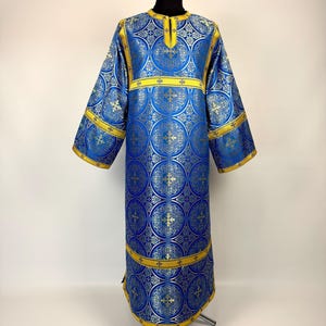 May include: A long, blue and gold liturgical vestment with a repeating circular pattern. The garment has long sleeves and gold trim around the edges and across the chest. The vestment is displayed on a black mannequin.